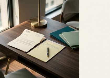 Desk setup by a window with a brass lamp, a yellow legal pad and pen, a printed letter, and blue-green folders stacked on a dark wooden desk.